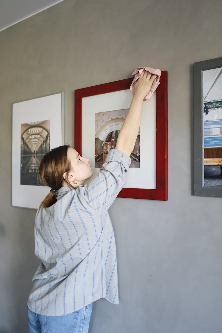 A woman cleans framed pictures on a wall, showcasing a tidy home environment.